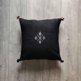 Black square cushion cover made of cotton with a decorative cross-shaped embroidered design and tasseled corners.