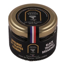 Black truffle flavored broken pieces in a glass jar with a gold lid. The jar is surrounded by leaves and soil, indicating the natural origin of the truffles.