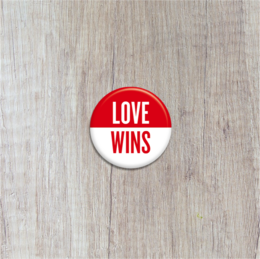 A round, red and white button with the text "LOVE WINS" printed on it. The button appears to be a brooch or pin that could be worn on clothing.