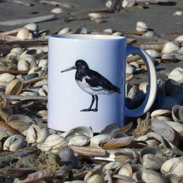 A white ceramic mug featuring a black and white illustration of an oystercatcher bird. The mug is surrounded by small pebbles and stones on a beach-like surface.