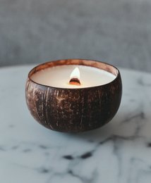 Scented coconut candle in a round, natural coconut shell container. The candle has a warm, tropical aroma and a simple, rustic design.
