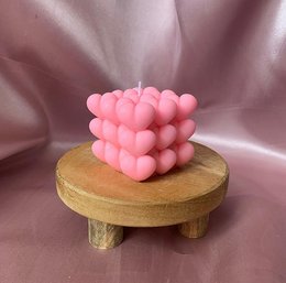 A round, pink candle with a cube-like shape, made of wax and sitting on a wooden stand.