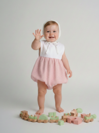  A pink and white baby romper with a ruffled skirt. The romper has short sleeves and a white top with a pink skirt. The baby is smiling and standing on a wooden floor with colorful building blocks.