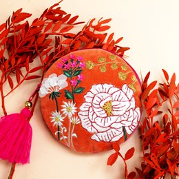 A round, orange purse with a floral design featuring a large white flower and smaller pink and yellow flowers. The purse has a tassel accent and is surrounded by vibrant red leaves.