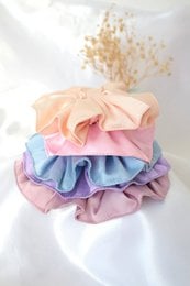 Handmade silk scrunchies set in pastel colors including pink, cream, lilac, and light blue. The scrunchies feature a soft, ruffled design and are suitable for various hair lengths.
