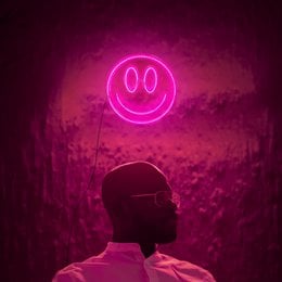 A round, pink neon smiley face lamp on a dark background, with a laptop and other lighting fixtures visible.