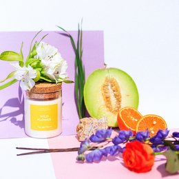A round, tall orange scented candle in a glass jar with a cork lid. The candle is surrounded by fresh flowers and citrus fruits, creating a vibrant and natural display.