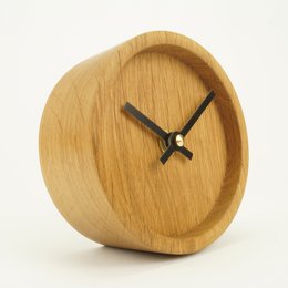 Cylindrical wooden table clock in oak, walnut, or black walnut finishes. Minimalist design with simple clock face.