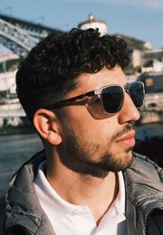 Black rectangular sunglasses with dark frames worn by a man with curly hair and a beard, standing in front of a cityscape.