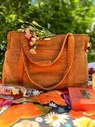 A large, orange corduroy tote bag with a floral pattern and a leather strap. The bag appears to be filled with various items, including flowers and leaves.