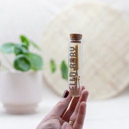 A glass tube filled with dried flowers and decorative elements, including a wooden label that reads "Hello Baby".