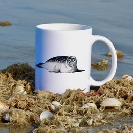 A white ceramic mug featuring a printed image of a seal resting on rocks near the ocean.
