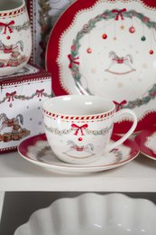  Porcelain cup and saucer set with holiday-themed design featuring a red and white color scheme, floral patterns, and a bow motif. Suitable for serving hot beverages.