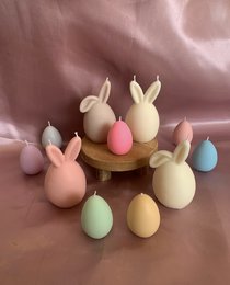 Pastel-colored Easter egg-shaped candles in various shades including white, pink, blue, orange, gray, and lavender. The candles are arranged on a wooden stand, creating a festive and decorative display.