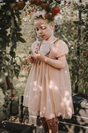 A delicate, handmade tulle dress in a soft peach color. The dress features a ruffled neckline and puff sleeves, creating a whimsical, feminine look. The dress is available in sizes 18-24 months to 9-10 years, making it suitable for a range of young girls.