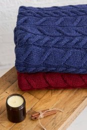 Navy blue and red cable knit throw blankets with a cozy, textured appearance. The throws feature a classic Aran cable pattern and appear to be made of soft, high-quality merino wool.