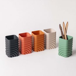 Wavy pencil/brush holder in ebony, cedar, and birch wood finishes. Cylindrical shape with textured, undulating surface. Suitable for organizing and storing writing utensils, makeup brushes, and other small office or vanity items.