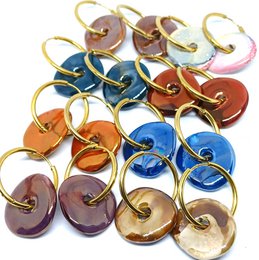 Large round hoop earrings made of ceramic in various vibrant colors including purple, orange, blue, and mauve. The earrings feature a donut-shaped design with a gold-tone metal frame.