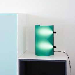  A modern, minimalist table lamp with a sleek, curved green glass shade and a black metal base. The lamp has a simple, elegant design that would complement a variety of interior styles.