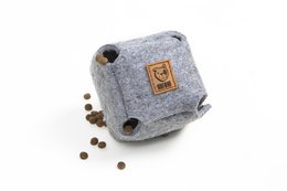 A round, gray pet toy with a paw print logo. The toy appears to be made of a soft, felt-like material and has a few coffee beans scattered around it.