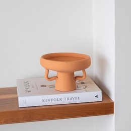  Handmade ceramic pottery dish in a warm orange color, featuring a round, elevated shape that would be suitable for serving small dishes or displaying decorative items.