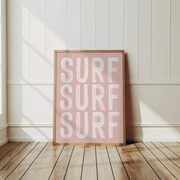 A pink and white poster with the text "SURF SURF SURF" repeated three times. The poster is framed and placed on a wooden floor.