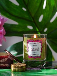 A tall, round glass candle with a pink label that reads "Yellow Neroli" sits on a green leaf background. The candle has a warm, flickering flame.
