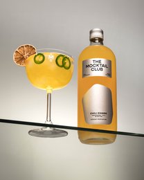 A 99cl bottle of Mocktail Chili Charm, a non-alcoholic beverage with a vibrant yellow color, garnished with a slice of lemon and green chili peppers. The bottle and glass display the product's branding and packaging.