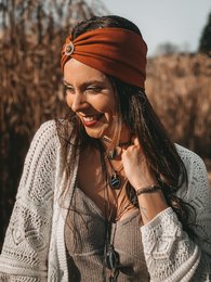 Rust-colored headband with a decorative brooch in the center, worn by a smiling woman in a cozy knit sweater.