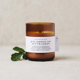 A tall, round, amber-colored glass candle with a label that reads "An evening on the Mediterranean". The candle is scented and comes in two sizes, 200 ml and 140 ml. The image also shows a green leaf, suggesting a natural or botanical scent.
