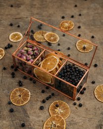 A small glass box containing various dried herbs, spices, and citrus slices. The box has a rustic, wooden frame and compartments for different ingredients.