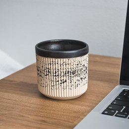 A ceramic mug with a textured, patterned surface in shades of beige and black. The mug has a simple, minimalist design with a round, cylindrical shape.