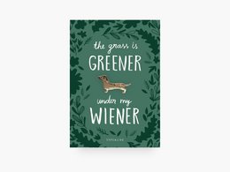 A green and brown brooch in the shape of a dachshund dog, with the text "The grass is greener when you're a wiener" on a leafy green background.