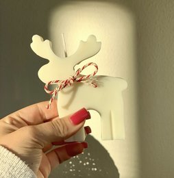 A round, tall white candle with a Rudolph the Red-Nosed Reindeer silhouette design and a red and white striped ribbon tied around it.