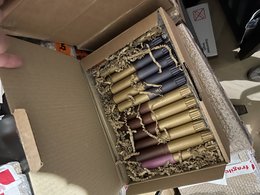 Assorted candles in a variety of colors and shapes, including tall, round, and decorative styles, packaged in a wooden box with shredded paper filler.