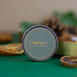 Spruce-scented soy candle in a round tin container. The candle has a green label with the word "Spruce" printed on it. The candle is surrounded by pine cones and dried orange slices, creating a cozy, natural atmosphere.