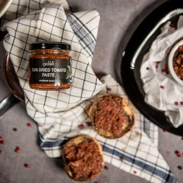 A jar of sun-dried tomato paste, a savory and flavorful spread made from sun-dried tomatoes. The jar is placed on a checkered cloth, along with some fresh tomatoes, suggesting the natural and homemade quality of the product.