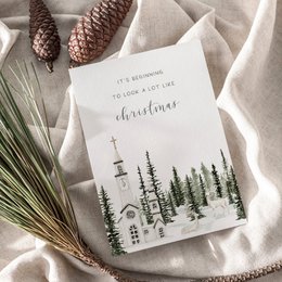 "Christmas card with winter forest scene and text 'It's beginning to look a lot like Christmas'. The card features a minimalist design with pine trees, stars, and other winter elements."
The image shows a Christmas card with a minimalist design featuring a winter forest scene. The card has a white background with a simple illustration of pine trees and other winter elements such as stars. The text on the card reads "It's beginning to look a lot like Christmas". The card appears to be made of a textured or structured paper material, and it is surrounded by other holiday-themed items like pinecones, dried orange slices, and a sprig of greenery.