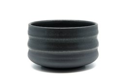 A round, black ceramic bowl with a matte finish and distinct horizontal ridges. The bowl contains a vibrant green powder, likely matcha tea.