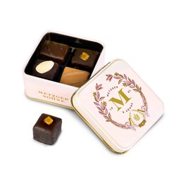  A pink tin containing petit gingerbread chocolates in various shapes and colors, including brown, yellow, and white. The tin is decorated with a floral wreath design.