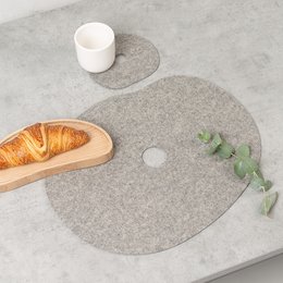 A round, gray wool felt placemat and coaster set. The placemat measures 36 x 29 cm and the coaster is 13 x 12 cm. The set features a minimalist, natural design suitable for everyday use.