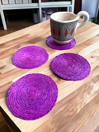 Set of 4 round woven palm straw coasters in vibrant purple color. Handmade from natural palm straw material, these coasters have a rustic, boho-chic style that is perfect for protecting surfaces from hot or cold drinks.