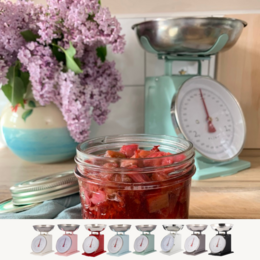 A cream-colored kitchen scale with a stainless steel bowl. The scale has a classic, retro design and can be used to weigh various food items. The scale is accompanied by a jar of what appears to be preserved or pickled strawberries.