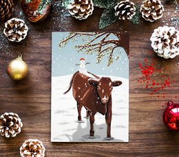 A snowy winter scene featuring a red poll cow standing in the snow, surrounded by pine cones and other festive decorations. The image evokes a cozy, rustic Christmas atmosphere.