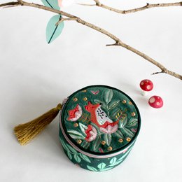A round, decorative jewelry box with a floral and fox design in shades of green, red, and gold. The box has a tassel accent and appears to be made of a sturdy material, likely wood or metal.