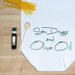 A white tea towel with the text "SEX, DRUGS AND OLIVE OIL" printed in green on a wooden surface, along with a bottle of olive oil and some spaghetti.