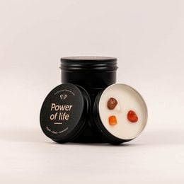 Scented candle in a black jar with the text "Power of life" on the label. The candle has a round, cylindrical shape and appears to be made of wax. The candle is surrounded by a few small, round objects that could be decorative elements or additional scents.