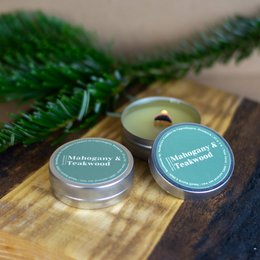  Mahogany and teakwood scented soy candles in round tin containers, with pine branches in the background.