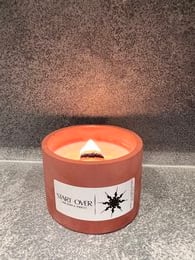 A small, round concrete candle with a warm, orange flame. The candle is labeled "START OVER" and has a simple, minimalist design.