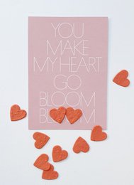 A pink card with the text "YOU MAKE MY HEART GO BLOOM BLOOM" and scattered red heart-shaped confetti. This love-themed postcard would be suitable for romantic occasions like Valentine's Day or anniversaries.